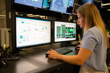 A woman working at a computer station with multiple monitors displaying technical data